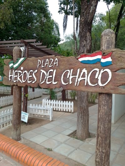 The image depicts a wooden sign that reads "Plaza Heroes del Chaco" in white lettering. The sign is decorated with flags that feature green, white, and blue stripes. The sign is supported by two wooden posts with a rounded top on the post to the right. Behind the sign is a white picket fence. A small sign written in Spanish is partially obscured by the post. The plaza is in Villa Hayes, Paraguay. The environment consists of greenery, trees, and a paved pathway.