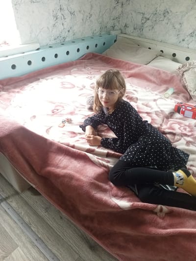 A young girl with blonde hair and glasses is lying on a bed covered with a pink blanket. She is wearing a black polka dot dress and black leggings, with yellow socks. The bed is in a room with light marble-patterned wallpaper. A light blue railing with circular cutouts runs along the top of the bed. There are several objects on the bed, including a small toy car, a baby bottle, and a keychain with a pink pig charm. The scene appears to be indoors, possibly a bedroom. The lighting suggests it is daytime. The girl's expression is neutral, and she is looking towards the camera. There is no visible text in the image. The location context is Buraly, Russia.