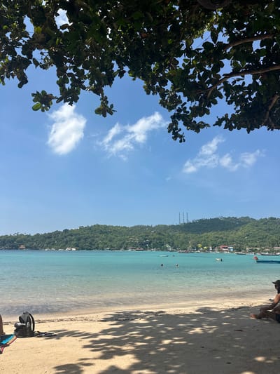 The image captures a vibrant, sunny tropical beach scene, likely in Koh Tao, Thailand, from a shaded vantage point. On the far left, the dense green foliage of a large tree dominates the foreground, providing natural framing and shade. Above the tree line, a wide expanse of clear blue sky with scattered, fluffy white clouds fills the upper half of the frame.
Stretching across the horizon in the mid-ground is a lush, green, tree-covered island or large hill, with scattered buildings and resorts visible along its base, nestled near the water. Three distinct antenna-like structures are also noticeable on the hillside.
Below the distant island, the calm, clear turquoise-blue water of the sea gently meets a light-colored sandy beach. Several individuals are seen swimming or wading in the refreshing water, enjoying the bright day. On the immediate right, the sandy beach is prominently featured, with strong, dark shadows from tree leaves cast across its surface, indicating direct overhead sunlight. A person is partially visible sitting in the shade on the sand, next to a dark bag and a colorful item of clothing or a towel. The clear sky and bright conditions suggest it is midday on a beautiful day, evoking a peaceful tropical vacation atmosphere. No visible text is present.