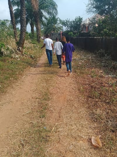 The image depicts an unpaved dirt pathway in Ufuma, Nigeria, during daylight hours. Four individuals are walking away from the camera, progressing along the path into the middle distance.

From left to right in the procession:
1.  A male individual wearing a white short-sleeved t-shirt and blue jeans.
2.  A male individual wearing a light grey long-sleeved shirt and dark trousers.
3.  A partially obscured individual, likely female, wearing a multi-colored patterned garment and dark hair.
4.  A male individual wearing a purple short-sleeved t-shirt and blue jeans.

All individuals appear to be walking casually.

The pathway is reddish-brown earth with a central, worn track. The immediate areas flanking the path are covered with sparse, dry, yellowish-brown grass interspersed with some green vegetation. A single light brown, dried leaf or seed pod is visible on the ground in the lower right foreground.

To the left of the path, several tall, slender palm trees with brown trunks stand amidst dense green and dry undergrowth. To the right of the path, a low, dark grey concrete or stone wall extends into the background. Beyond this wall and a dense cluster of green trees and bushes, the upper portion and grey roof of a light-colored building are visible against the sky. The sky is partially visible, appearing bright with some haze or light cloud cover.