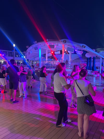 This is a photograph of a nighttime outdoor party or dance event on a wooden deck, likely aboard a cruise ship or at a resort, given the presence of deck chairs and umbrellas in the background. A group of people are dancing and socializing under colored lights, with beams of blue and red light illuminating the scene. The overall atmosphere is festive. The image does not provide any information to suggest a location in Salobreña, Spain.