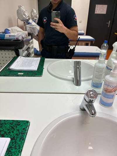 A man in a uniform holds a smartphone, presumably taking a selfie, in front of a white sink and counter. On the counter are soap bottles, a small water bottle, and a chrome faucet. A clipboard with papers is attached to the cabinet below the sink. Next to the counter is a cart with medical supplies. A small portion of a green folder with black spots is visible on the right. The image is taken at an unusual angle.