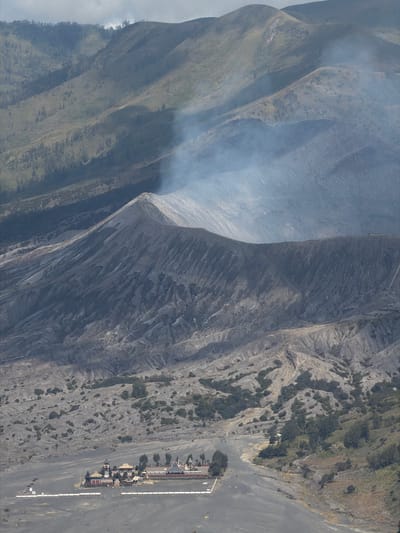 The image captures an aerial view of the Pura Luhur Potong Temple, located in Cemorolawang, Indonesia, set against the backdrop of volcanic mountains. Smoke plumes rise from a distant peak, indicating volcanic activity. In the foreground, the temple complex is visible, characterized by its distinct architectural style with red and dark-colored roofs and ornate details. Several people can be seen walking around the temple grounds and on the adjacent sandy terrain. The surrounding environment consists of barren, ash-covered slopes interspersed with sparse vegetation. The overall scene depicts a stark and dramatic landscape, with the temple serving as a point of human habitation within the active volcanic region.
