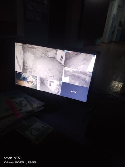 The image shows a computer monitor displaying a multi-camera surveillance feed. The monitor is positioned on a desk with papers and a disability access symbol visible in the foreground. The surveillance feed shows several different camera views of what appear to be outdoor and indoor areas of a building, possibly a school or office complex, with timestamps indicating the date "07-03-2026" and time "23:34:21". Some of the indoor views show hallways and rooms with furniture. The lighting in the image is dim, with light primarily emanating from the monitor. The overall setting suggests a security monitoring station. There are no identifiable people or specific locations within the city of Мәскеу, Kazakhstan, visible or suggested by the image content itself.