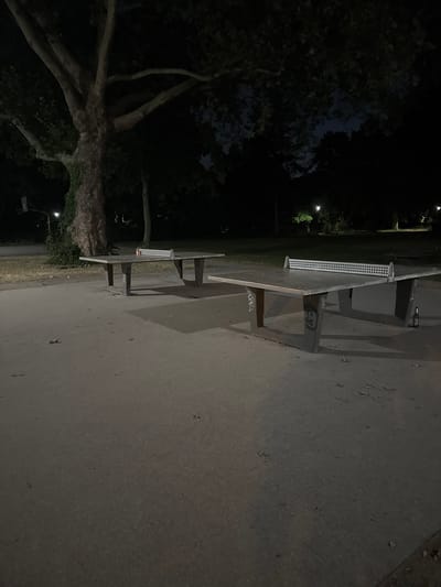 This is a nighttime photograph of two outdoor concrete ping pong tables with nets. The tables are located on a paved area in a park, with a large tree visible to the left. In the background, there are more trees and lampposts providing artificial light. A bottle of beer is placed on the ground next to the right ping pong table. The scene appears to be in the Rheinpark in Duisburg, Germany, a well-known public space. There are no people visible in the image.