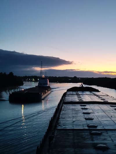 The image shows two barges navigating a waterway at dusk. The photograph is likely taken from the deck of one of the barges, showing the other barge in the distance. The sky is a mix of blues and oranges, indicating either sunrise or sunset. The waterway is lined with trees on both sides. Light reflects off the surface of the water. The location is speculated as the city of Annensky Most, Russia.