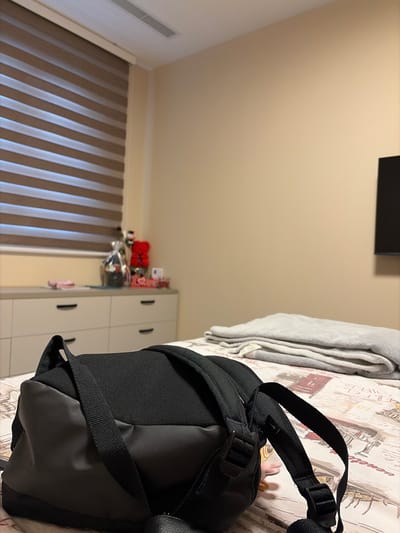 This media file captures a close-up view of a black backpack placed on a bed. The backpack is the primary subject, positioned in the foreground, with its straps and textured fabric clearly visible. The bedspread beneath it has a vintage, patterned design, featuring illustrations that appear to be from a historical context, possibly related to cityscapes or architectural elements.

In the background, a set of white drawers with black handles is visible, suggesting a bedroom or living space. Above the drawers, on a shelf, are several items, including what looks like a bouquet of red flowers and other decorative objects, some of which are wrapped. To the left of the drawers, a window with horizontal blinds is present, their alternating light and dark stripes creating a rhythmic pattern. The wall is painted a neutral, light color. A portion of a gray blanket or clothing item is draped on the bed, to the left of the backpack. In the upper left corner, a dark rectangular object is mounted on the wall, possibly a television or a frame. The overall setting appears to be indoors, likely in a residential room. No people are visible in the media file. The lighting suggests it could be daytime, but the exact time of day is not discernible. The location context is Brestnik, Bulgaria. There is no visible text in the image.
