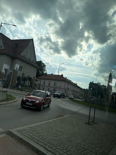 A red Dacia Sandero, license plate KLI 86051, turns right onto a paved road in Limanowa, Poland. The car is in the foreground, facing away from the viewer. In the background, a building with a mansard roof and stone facade is visible, with an advertisement poster attached to its side. Other buildings line the street, and a row of trees can be seen behind them. The sky is filled with dramatic, cloudy formations. Several birds are scattered throughout the sky. The overall scene depicts an urban streetscape under a dynamic sky.