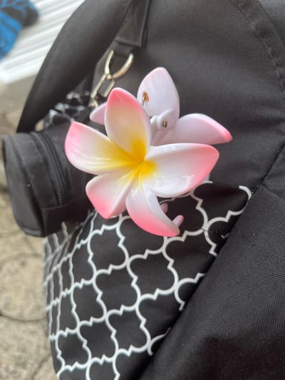 A close-up, angled outdoor shot focuses on a pink and yellow plumeria hair clip attached to the side of a black bag. The bag features a black and white quatrefoil pattern on a portion of its surface and has a zippered pocket on its left side. The background is out of focus, showing a hint of a white slatted surface and some blue material on the upper left, and a textured, light-colored surface, possibly pavement or stone, in the lower left. The lighting suggests it is daytime, with diffused light. The scene is not specifically identifiable as being in Oberammergau, Germany.