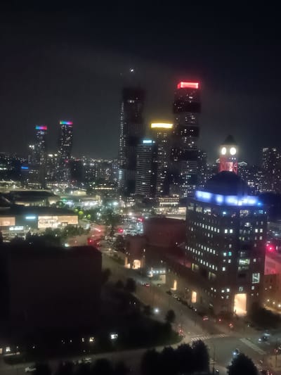 This is a nighttime aerial view of the city of Mississauga, Canada, specifically the area around Square One Shopping Centre. The image shows a cityscape dominated by illuminated skyscrapers and buildings. In the foreground, a darker, larger building is visible. Further into the background, several prominent skyscrapers stand out, including two towers with colorful, illuminated tops and another with a red illuminated section. The cityscape is dotted with numerous lit windows, indicating occupied buildings. Roads with vehicle headlights and taillights are visible, suggesting active traffic. The overall scene is one of a vibrant, illuminated urban environment at night.