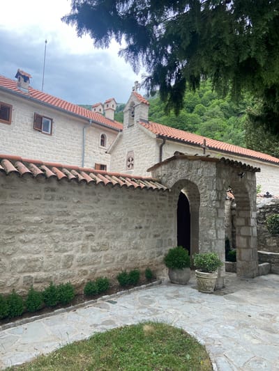 The image depicts a stone building with a red tile roof, located in Podličak, Montenegro. The building has an arched entryway with stone pillars. A stone pathway leads to the entrance. A planter with a green plant sits near the entryway. A strip of small green bushes lines the side of the building next to the pathway. In the background, trees and other buildings are visible. The sky is overcast.