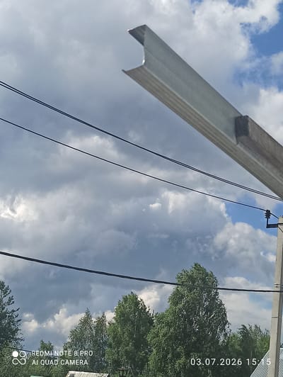 The image depicts an outdoor scene with a cloudy sky overhead. In the foreground, a metallic beam is angled from the upper right to the lower left, supported by a wooden block. Several dark power lines are strung horizontally across the frame. Below the power lines, lush green trees are visible, with some appearing taller and fuller than others. In the lower left corner, text overlays indicate "REDMI NOTE 8 PRO AI QUAD CAMERA", and in the lower right corner, a date and time stamp reads "03.07.2026 15:15". A concrete pole with electrical insulators is present on the right side of the image. The overall scene appears to be a somewhat overcast day in a semi-rural or suburban environment, possibly Sovetskiy, Russia, given the location tag. There are no people or specific actions depicted.