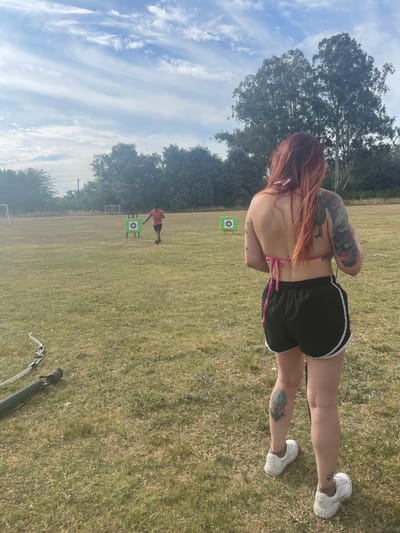 An outdoor scene shows a woman with reddish hair, tattooed arms, and white sneakers, posing with her back to the camera. She wears a pink top and black shorts with white trim. Further away, a person in red stands near archery targets on a grassy field with a few trees and a blue sky with clouds. An archery bow and quiver are on the ground near the bottom of the image.