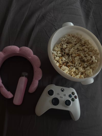 This overhead shot shows a white bowl filled with popcorn, a white Xbox controller, and a pink fuzzy headband with a pink vape device resting on it. The items are arranged on a dark gray, wrinkled fabric surface, possibly a bed or sofa. The lighting is dim, suggesting it might be nighttime or an indoor setting with low light. The overall scene evokes a sense of relaxation and entertainment, implying someone is preparing for a gaming session or a movie night. There is no visible text in the image. The location is identified as Hausach, Germany.