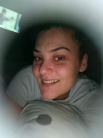 The image, purportedly captured in Iskrets, Bulgaria, presents a single individual, identified as an adult female, positioned in a dimly lit indoor environment. The subject is centrally framed, facing directly towards the viewer with a slight smile, revealing her teeth. Her eyes are brown, and her eyebrows are dark and well-defined. Visible redness is present on her cheeks and beneath her eyes. Her dark hair appears to be pulled back, with some wisps at the hairline. She is wearing a light-colored t-shirt, possibly pale grey or light blue.
Her head rests on a light-colored, soft object, likely a pillow, which exhibits a distinct dark, circular stain or pattern positioned near the bottom center of the frame. In the upper left background, a rectangular screen, possibly a digital device, is visible at an angle. The screen displays text written in Cyrillic script; partially legible words include "ПРИ УВОРА" and "ИМУСКУЛИ". The overall lighting of the scene is low, with a soft, diffused light source illuminating the subject's face, creating a halo effect around her. The immediate background behind the subject is dark and indistinct.
