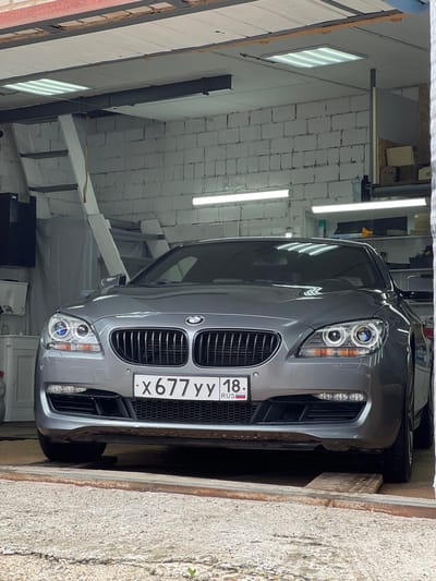 The image shows the front of a grey BMW parked inside a garage or workshop. The vehicle is positioned on a slightly raised platform, possibly for repair or detailing. The workshop appears to be constructed with white brick walls and has fluorescent lighting fixtures on the ceiling. There are shelves visible in the background with various items, including boxes and bottles. A ladder-like structure is also present on the left side of the image. The license plate of the car is clearly visible and reads "x677yy 18 RUS". There are no people or animals visible in the image. The overall scene is static and appears to be a snapshot of a vehicle in a professional service environment.