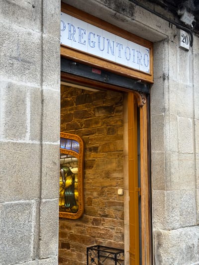 A street-level exterior shot shows the entrance to a business in Santiago de Compostela, Spain. The business name, "PREGUNTOIRO", is displayed in blue lettering on white tiles framed by wood above the entrance. To the right of the entrance, a sign with the number "20" is affixed to the stone wall. Inside the entrance, a stained-glass window with the text "DE LA VIDA" is visible. The architecture features rough-hewn stone blocks on the exterior walls.