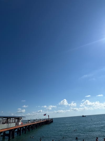A long pier extends into the calm, blue water of the Black Sea under a clear, bright sky in Adlerskiy, Russia. The pier is lined with a railing and has a building structure on the left side, with solar panels on its roof. People are visible on the pier and swimming in the water. A Russian flag is flying on a pole on the pier, and a small boat is visible in the distance on the horizon. The sun is shining brightly, casting a slight glare on the water and sky. The overall scene depicts a pleasant day at the seaside.