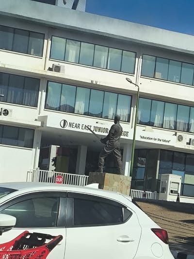 An outdoor daylight scene depicts the entrance of the "NEAR EAST JUNIOR COLLEGE" in Kızılbaş, Northern Cyprus / Cyprus. The building features a light-colored facade with multiple floors and numerous reflective, rectangular windows. "NEAR EAST JUNIOR COLLEGE" is prominently displayed above the main entrance, accompanied by a circular logo. To the right, a smaller sign reads "education for the future."
In front of the building, a dark-colored, possibly bronze, statue of a male figure stands on a stone-clad pedestal. The figure is depicted in a dynamic, athletic pose, holding a long implement, resembling a sword or bat, with both hands, positioned as if about to swing. The statue's back is oriented towards the viewer. A green street light pole is situated to the right of the statue. White metal railings separate the immediate foreground from the college entrance area. A red octagonal sign with the word "DUR" in white lettering is visible behind a white car.
A white hatchback car, with tinted rear passenger windows, occupies the lower portion of the image, obscuring parts of the statue's base and the building's ground level. A red plastic shopping cart is partially visible in the bottom left corner of the frame. The sky above is clear blue, indicating a sunny day.