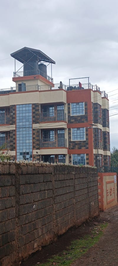 A multi-story building behind a high stone wall is captured on a cloudy day. The building has a water tank structure on its roof, with a person visible on the top floor balcony. The building is constructed of brown stone, orange accents, and large reflective windows. The wall is made of dark stones and extends along the foreground, partially obscuring the building. A portion of a sidewalk is visible in the foreground, with sparse greenery.