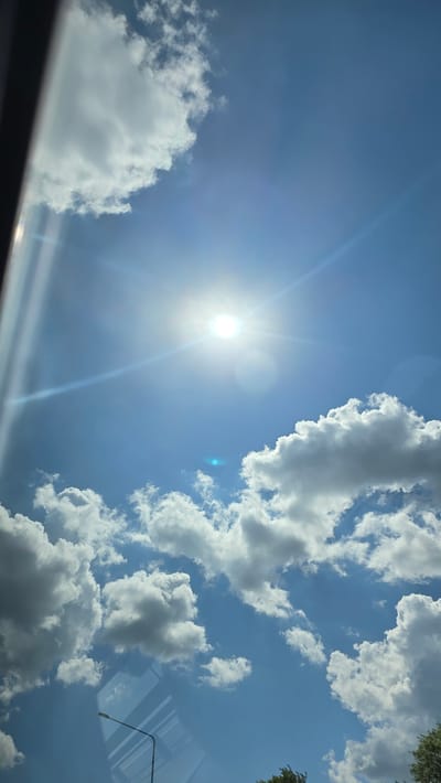 A vertical, full-frame photograph captures a bright, sunny sky with scattered white clouds. The sun is prominently positioned in the upper center of the frame, emitting strong rays that create lens flare and a visible blue light artifact. A dark, vertical element, possibly a window frame or obstruction, is visible on the left edge of the image. In the lower portion of the frame, a street lamp with a curved arm is visible, and reflections of what appear to be windows or a ceiling are superimposed on the sky. Some green foliage can be seen at the bottom right corner, suggesting an outdoor environment. The scene is consistent with a view from a vehicle or indoor location looking outwards on a clear day. The location is specified as Tolbazy, Russia.