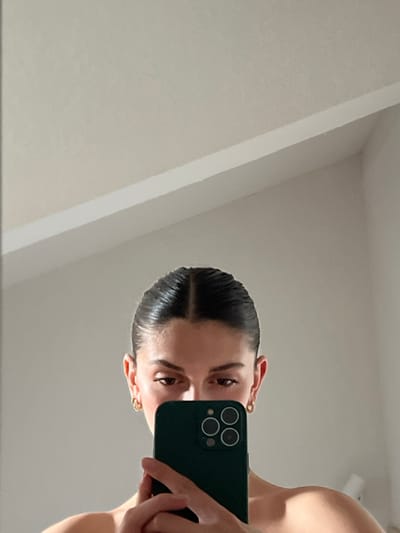 The image shows a woman holding a black smartphone with a dark green case in front of her face. She is looking at the phone's screen. She has dark brown hair slicked back and is wearing gold hoop earrings. She is nude from the chest up, only her face and upper torso are visible in the image. She appears to be indoors. The background is white, with a white ceiling and a white wall with a slight shadow. It is difficult to determine the location based on the image. The latitude and longitude coordinates provided do not match the location in the image.