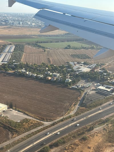 The image is an aerial view, taken from an airplane, of a rural and semi-urban landscape in Kfar Truman, Israel. The left side of the frame is dominated by the wing of the airplane, which is light blue and white with visible panel lines and markings. The landscape below is a patchwork of agricultural fields, some appearing fallow with dark brown soil, and others showing rows of crops, possibly green vegetables or trees.
A small village or settlement is nestled between the fields, characterized by numerous white and light-colored houses with red-tiled roofs, surrounded by lush green trees. Adjacent to this settlement is a multi-lane highway, on which several white cars are visible, suggesting moderate traffic. The highway runs through dry, grassy terrain. Further to the left, a denser urban area with taller buildings can be seen in the distance, shrouded in a light haze.
The scene appears to be captured during the daytime, likely in the late morning or early afternoon, given the bright sunlight casting shadows. The weather seems clear and dry, as indicated by the crisp visibility and the appearance of the landscape. The overall impression is one of a developed agricultural region with expanding settlements and infrastructure. There is no visible text in the image.