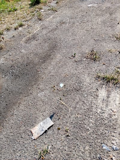 The image shows a close-up of a paved surface with scattered debris. In the foreground, a small, flattened object that appears to be a used condom wrapper lies on the asphalt. Beyond this, the ground is a mix of rough asphalt and patches of dry grass and weeds. The lighting suggests it is daytime. The context provided indicates this scene is located in the city of Bytom, Poland, implying an urban or suburban environment. There are no people or visible actions occurring in the image.