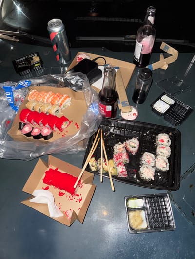 This media file depicts a messy scene of leftover sushi and drinks on a dark, flat surface, likely a table or countertop, with hints of a gritty or industrial background. The primary subjects are various containers of sushi, empty and partially full bottles of wine, a can of soda, and disposable chopsticks.

The sushi is presented in several black plastic containers and a brown cardboard box. One container holds several pieces of sushi rolls with white rice and what appears to be imitation crab meat. Another container has rolls topped with red roe and sesame seeds. A brown cardboard box contains a selection of sushi rolls, some topped with orange shrimp, others with white rice and sesame seeds, and some covered in red roe. A smaller brown cardboard box contains a portion of red roe, likely from a sushi dish. There are also small plastic containers holding soy sauce and wasabi.

Several glass bottles, some containing red wine, are scattered across the scene, along with a silver soda can. One wine bottle is mostly full, while another appears to be empty or nearly so. A brown paper band with blue text is also visible, possibly from a food item.

The overall impression is that of a meal that has concluded, with the remnants left behind. The lighting is dim, suggesting it could be nighttime or an indoor setting with low light. There are no people visible in the image. The presence of disposable containers and the disarray point towards a casual or takeout meal.

Visible text includes "NON STOP" on the soda can, and partial text on the wine bottle labels and the brown paper band. The location context of Bavoriv, Ukraine, does not offer specific visual cues within the image itself, but helps contextualize the scene as a snapshot of daily life in that region.