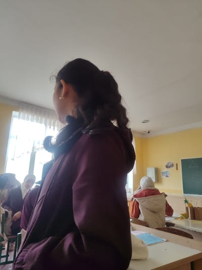 The image depicts an indoor scene, identified as a classroom or educational setting in Rishtan, Uzbekistan. The primary subject is a young individual, likely female, observed from the upper back and right side, positioned in the foreground. This individual has dark, curly hair pulled back and wears a dark purple hooded jacket. A small, light-colored stud earring is visible in their right ear. They are seated at a light-colored wooden desk, upon which a light blue booklet and a white object are present.

In the mid-ground, to the left of the primary subject, another individual, possibly female, is partially visible, wearing a white head covering and a dark garment, facing towards the left. Further into the background, another person is seated facing away from the camera, wearing an orange top and a light-colored quilted vest.

The room features yellow walls in the upper sections and a white ceiling. A large window with white sheer curtains is on the left, admitting significant natural light. On the far wall, a dark green chalkboard displays a white triangle and a white circle. Below the chalkboard, several light-colored paper bags with star decorations and green ornaments are visible. A white electrical panel is mounted on the yellow wall. A small, dark security camera is affixed to the white ceiling in the background. Colorful papers or drawings are taped to the yellow wall near the chalkboard.