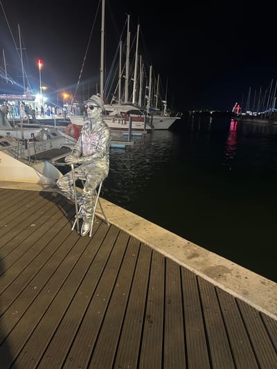A silver-painted street performer sits on a folding stool on a wooden boardwalk at night in Agde, France, near the marina. The performer is dressed in a shimmering silver outfit, complete with a hat and sunglasses, and holds a silver cane. Behind the performer, several sailboats are moored in the dark water, their masts reaching into the night sky. In the distance, a Ferris wheel with red lights is visible, suggesting a fair or festival. The boardwalk is made of dark wooden planks, and a concrete barrier separates it from the water. Several other boats and a lamppost are also visible in the background, along with a crowd of people in the distance.