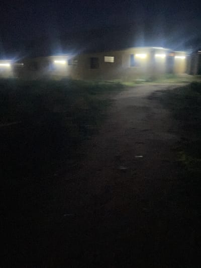 The image depicts a dark, outdoor scene at night, likely in Safari, Nigeria. In the background, several single-story structures are visible, illuminated by bright interior lights shining through windows and doorways. The buildings appear to be made of a light-colored material, possibly concrete or stucco, and are arranged in a row or cluster. A dirt path or road leads towards these structures from the foreground. The ground in the foreground is dark and appears to be unpaved, with some patches of grass or vegetation visible. The overall impression is of a residential or communal area at night, with artificial lighting providing the primary illumination. There are no people visible in the image.