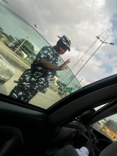 The image depicts an outdoor scene in Eneka, Nigeria, viewed from within a vehicle. A male individual, identified as a Nigerian police officer by his camouflage uniform featuring the "NIGERIA POLICE" emblem on the left sleeve and a matching boonie hat, stands outside the vehicle. He is positioned towards the front-left side of the vehicle, with his right hand raised, palm open, facing the interior. A black baton is visible secured to his left hip. The officer displays a slight smile. The vehicle's dark interior, including the sunroof frame, part of a seatbelt, and the driver's hands on a steering wheel, wearing a white shirt, occupies the lower portion of the frame. In the background, a paved road surface is visible with various vehicles, including a light-colored SUV and a yellow auto-rickshaw (Keke Napep). Several structures with green roofs or awnings, along with tall utility poles and a large tower-like structure, are present under a partly cloudy sky.