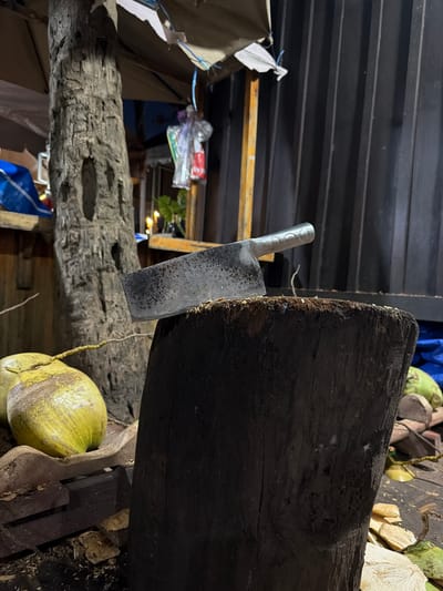 A cleaver rests on a dark wooden chopping block, positioned in front of a weathered tree trunk at an outdoor market in Luang Prabang, Laos. Several coconuts are visible, with one yellow coconut in the foreground and others in the background. A corrugated metal wall forms the backdrop, with some overhead shelter structures and decorative items hanging in the upper portion of the image. The scene appears to be set up for selling coconuts, likely in the evening due to the lighting.