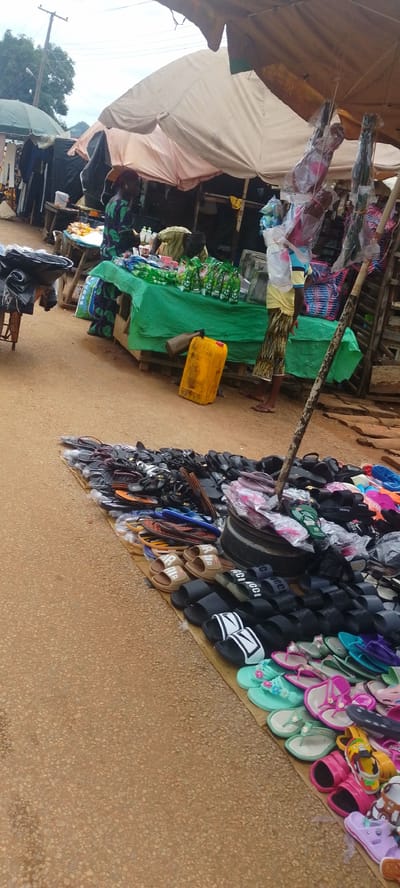 This is a photograph taken in Baissa, Nigeria, depicting a bustling outdoor market scene. The foreground is dominated by a large display of footwear, primarily flip-flops and sandals, spread out on the ground. Various colors and styles are visible, suggesting a diverse selection for customers.

In the midground, several stalls are set up. One stall, covered with a green cloth, displays bottles of what appears to be beverages, along with other packaged goods. Behind this stall, a person in a patterned dress is partially visible. To the left of this stall, another person in a green and black patterned garment stands near a table with items. Further back, clothing items hang on display.

The setting appears to be a roadside market, with a dirt or gravel road surface in the foreground. The sky is overcast, suggesting it might be daytime with cloudy weather. Overhead, a large, light-colored tarp provides shade for some of the stalls, supported by wooden poles. The overall impression is one of a vibrant local marketplace, likely busy with shoppers and vendors. There is no visible text in the image.