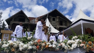 A group of men stand on a platform decorated with white and orange flowers. The men are wearing white robes, and one of the men holds a sword. The platform is in front of a building with a white tent and a wooden structure behind it. The location is Rukoko, Democratic Republic of the Congo. The coordinates are latitude: -1.624, longitude: 29.258. There is a blue sky with white clouds in the background.