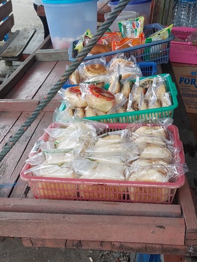 In Parit Baru, Indonesia, a close-up shot captures a rustic wooden market stall overflowing with baked goods. In the foreground, two red plastic baskets are filled with individually wrapped buns. The top basket, a vibrant green, contains more buns also individually wrapped. Behind these, a blue plastic basket holds an assortment of packaged snacks, some with colorful wrappers. In the background, a large blue translucent plastic container is visible, and a pink plastic basket sits on a cardboard box. The overall scene suggests a street-side vendor operation.