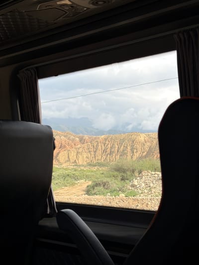The image is taken from inside a vehicle, likely a bus, looking out of the window at a natural landscape. The foreground shows the backs of two dark seats, with one in the foreground on the right and another in the foreground on the left. Between the seats, a window reveals a view of dry, rugged terrain with layered rock formations in shades of orange and yellow. Green vegetation, including bushes and grass, grows in the lower parts of the landscape and along a dirt road that winds through the scene. In the background, distant mountains are visible, partially obscured by clouds. The sky is overcast with a soft, diffused light. The scene appears to be located in the vicinity of Tong, Kyrgyzstan, known for its dramatic canyons and mountainous scenery.