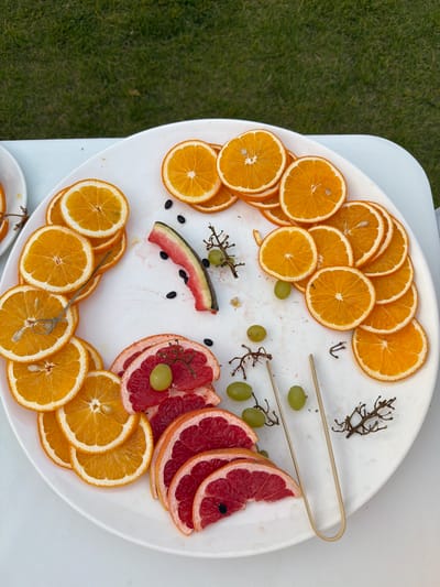 A overhead shot displays a white platter filled with an assortment of sliced citrus fruits and grapes, set on a white surface with a patch of green grass to the left. The platter is generously filled with thin, circular slices of dried oranges, fanned out in a decorative pattern around the edges. Interspersed among the oranges are several ruby-red slices of grapefruit, also arranged artfully. A few small, green grapes are scattered across the platter, some adorned with thin, twig-like decorations. In the center, a thin slice of watermelon with its distinctive rind and red flesh is visible, accompanied by a few dark, oval-shaped seeds. Two pairs of wooden tongs rest on the platter, suggesting the fruit is meant to be served or picked. The lighting suggests it is daytime, possibly outdoors, given the presence of the grass. The overall impression is that of a refreshing and visually appealing fruit presentation, likely for a gathering or event. No text is visible in the image.