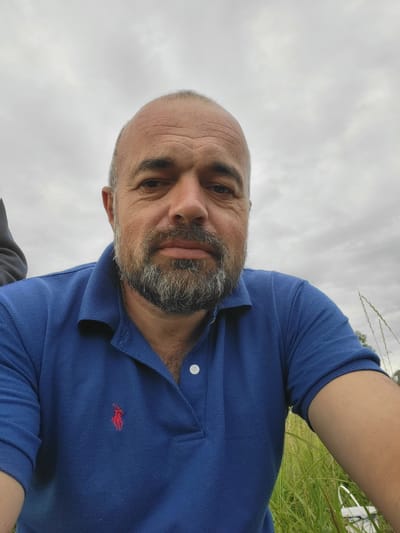A man with a bald head and a gray beard is captured in a close-up, outdoor selfie. He is wearing a blue polo shirt with a red logo on the left chest. The background consists of a cloudy sky and tall green grass, suggesting a rural or park-like setting. The image was taken in Hetmanivka, Ukraine.