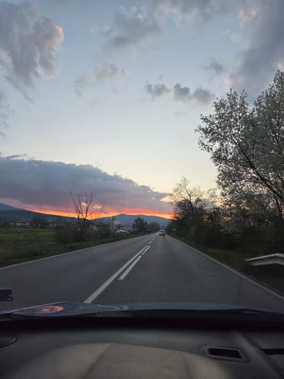The image shows a road in Piperkov Chiflik, Bulgaria at sunset. The view is from inside a moving vehicle, looking forward. The road has a white dividing line and is flanked by green vegetation on either side. On the left, the sky is a mix of grey clouds and orange hues from the setting sun. A line of trees and bare branches silhouette against the sky. In the distance, other vehicles can be seen on the road. Part of the interior of the vehicle is visible on the right side of the frame.