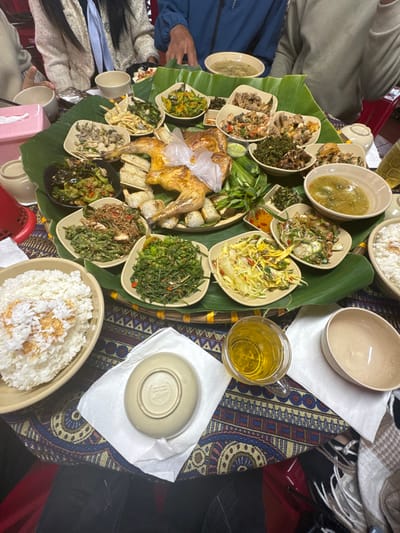 A table laden with a feast. A large, round woven platter is covered in banana leaves, displaying a roasted chicken, tofu, and various dishes of vegetables. The food is served in small bowls and plates. Steaming bowls of rice sit nearby. A clear mug of tea and empty bowls complete the setting. The tablecloth features a colorful, patterned design. Several individuals are seated around the table, seemingly ready to enjoy the meal.
