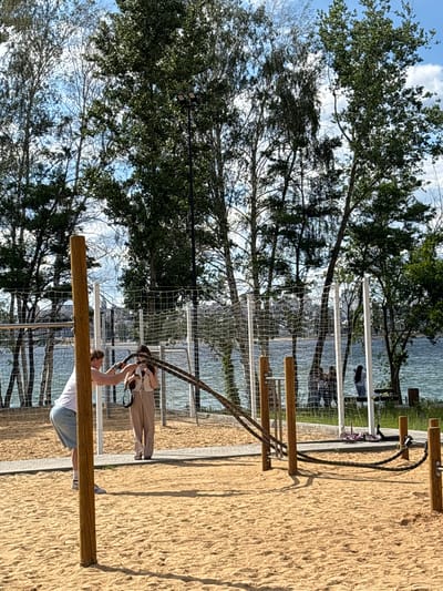 The image depicts an outdoor fitness area with sand ground. In the foreground, a man in a white t-shirt and denim shorts is performing battle rope exercises. To his right, a woman in a beige outfit stands nearby. Behind them, a net encloses the fitness area. In the background, a body of water is visible, with trees and buildings on the opposite shore. Several other individuals are present in the background, some near the water and others near a picnic table. The scene is set on a sunny day with a blue sky and scattered clouds. This location is in Novaya Usman, Russia.