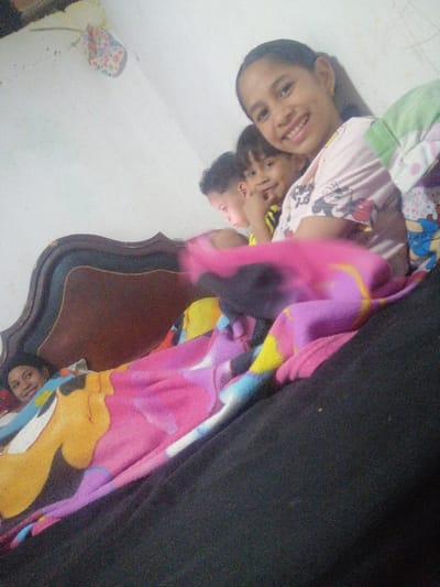 This image depicts an indoor scene, identified as being in Soacha ciudad, Colombia, likely a bedroom. Four individuals are present in a bed. In the foreground, a female with dark hair smiles directly at the camera, wearing a light-colored t-shirt with what appears to be Mickey Mouse graphics. Behind her and to the left, a younger child with dark hair is partially visible, also looking towards the camera with a finger near their mouth, wearing a yellow and black striped top. Further left, a third child, a boy with short dark hair, is visible, looking downwards. On the far left, partially obscured by a colorful blanket and the ornate dark wooden headboard, another individual, possibly an adult, is seen lying down, looking towards the camera with a slight smile. The individuals are covered by multiple blankets, including a prominent pink, purple, yellow, and blue blanket with patterned designs, and another light green and white blanket. The background consists of a plain white wall. An object, possibly a floral-patterned fabric decoration, is partially visible hanging on the upper left wall. The image exhibits a slightly soft focus, suggesting a casual, handheld photograph of a relaxed indoor gathering.