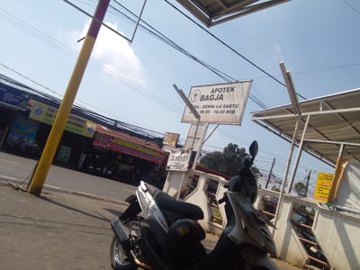 The image depicts a street scene in Cianjur, Indonesia, on a bright, sunny day. In the foreground, two motorcycles are parked, one silver and one black. The silver motorcycle, partially visible on the left, has a "Yamaha" logo and a "Mio" decal. A black helmet rests on its seat. The black motorcycle, on the right, is mostly obscured.
Behind the motorcycles, a sidewalk runs along a street lined with various businesses. A prominent yellow and red utility pole stands on the left side of the frame. Several signs are visible, including "APOTEK BAGJA" which indicates a pharmacy. Another sign advertises "PRIMO MOTOR" specializing in body parts and motor repair. Faded signage for laundry services is also present, mentioning "TIK LAUNDRY" and a price of "RP 7.000/KILO".
Overhead, a network of power lines crisscrosses the sky. The sky is blue with scattered white clouds. The architecture suggests a commercial area with modest buildings. The overall impression is one of a typical urban environment in a Southeast Asian city.
