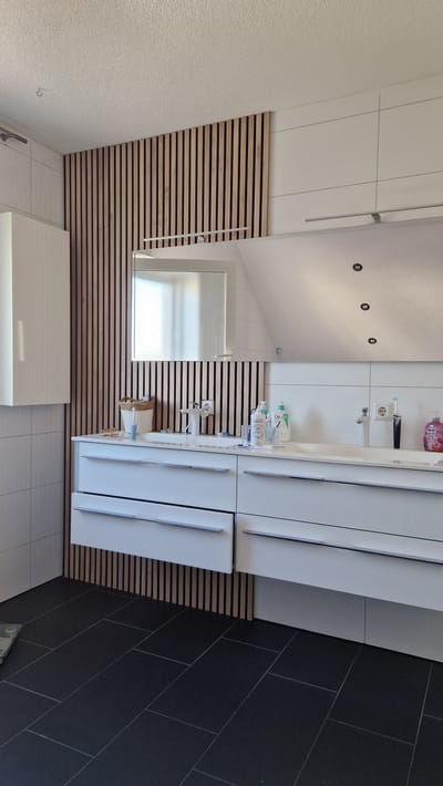 The image depicts a modern bathroom with a sloped ceiling, likely in a house in Ichenhausen, Germany. The primary subjects are various bathroom fixtures and furniture. A white vanity with multiple drawers is prominent on the right side of the frame, topped with a white countertop. On the countertop, several bottles of toiletries and personal care items are arranged, including what appears to be CeraVe products, a hand soap dispenser, and toothbrushes. A white sink with a chrome faucet is integrated into the vanity. Above the vanity, a large mirror reflects the ceiling lights and part of the room.

To the left of the vanity, a section of the wall is covered with horizontally slatted wooden panels, creating a natural and textured contrast with the clean white surfaces. A frosted glass window with horizontal blinds is set into the wooden paneling. Further to the left, the wall has a rough, textured white finish. On the floor, dark, rectangular tiles are laid in a staggered pattern. In the foreground, a white cabinet or countertop is visible.

The scene appears to be an interior shot of a well-lit bathroom, suggesting it's daytime. There are no people present, and the atmosphere is one of clean, functional, and contemporary design. No specific events are taking place; it is a static view of the room. Notable details include the sloped ceiling, the combination of wood and white finishes, and the dark tiled floor. No discernible text is visible on the objects within the image.