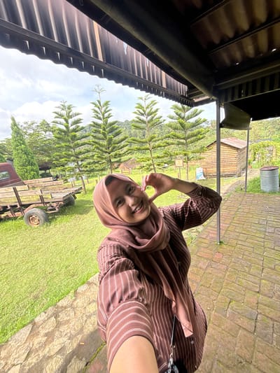 A smiling woman wearing a headscarf and striped brown shirt gestures with her hand, forming a heart shape near her face. She is standing in a green grassy area with trees in the background. Part of a wooden structure and a metal roof are visible on the left. A wooden wagon sits in the grass near her. In the distance, a small wooden building and a metal bucket-like container are visible. The ground in the foreground appears to be paved with bricks.