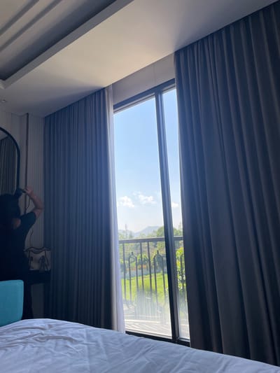 The image displays an interior view of a room, likely a hotel or guesthouse, in Kayuambon, Indonesia, looking out through a large, dark-framed glass window or door. In the immediate foreground, a white, rumpled bedsheet is visible. To the left, a person with dark hair, wearing a dark t-shirt, stands with their back to the camera, holding a black device (possibly a camera or smartphone) up to capture the outdoor view. Behind the person, a dark-framed arched mirror, a dark piece of furniture, and a light-colored tote bag with a patterned item are visible against a vertically paneled wall. A bright blue object, possibly a furniture accent or headboard, is situated in the lower-left corner.

The interior space features a modern white ceiling with stepped architectural detailing and recessed lighting channels. Two sets of pleated curtains frame the window: a lighter grey-blue set partially drawn on the left, and a darker grey set mostly drawn on the right.

Beyond the window, a clear blue sky with scattered white cumulus clouds is prominent. In the mid-distance, green hills or mountains are visible under the bright sky. Immediately outside the window, a dark, ornamental metal railing indicates a balcony or patio. Below the railing, lush green trees, foliage, and a patch of grass are present. A slender, distant communication tower or similar structure is faintly visible on a hillside to the far right. The scene is brightly illuminated by natural light entering through the window.