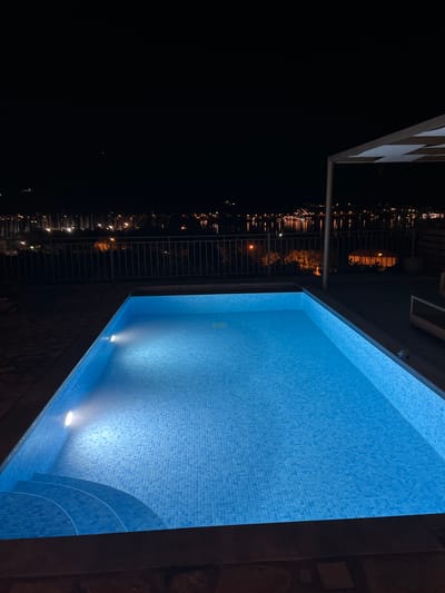 A nighttime aerial view captures a rectangular swimming pool, illuminated by underwater lights, in the Municipal Unit of Ellomenos, Greece. The pool is situated on a paved patio with a dark grey border. Steps descend into the pool on the left side. Beyond the pool, a metal railing separates the patio from a landscape featuring a body of water with numerous sailboats docked. The distant shoreline is dotted with lights from a town or city, suggesting an urban or suburban setting. A white-tented structure with a checkered pattern is visible in the upper right corner of the frame. The sky is dark and clear.