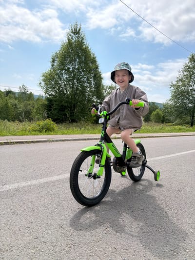 A young boy with a bucket hat and a grey jacket rides a bright green bicycle with training wheels on an asphalt road. The boy is smiling and looking towards the camera. The background shows a line of trees and hills under a partly cloudy sky. The scene appears to be in a rural or suburban area, possibly near the city of Kuldur, Russia.